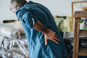 man with back pain in a workspace surrounded by various materials related to a home project experiencing discomfort lifting nine items