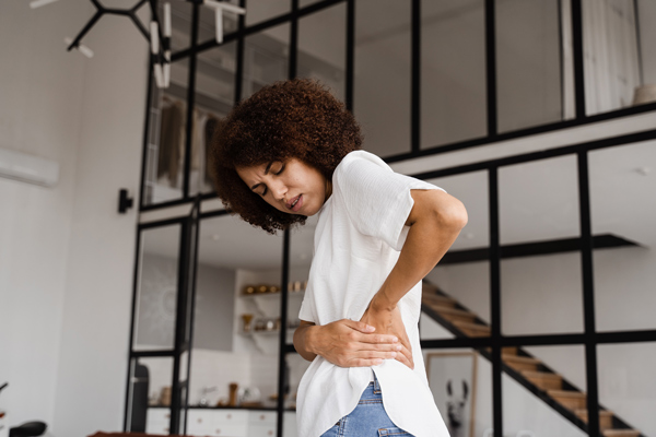 A woman in a bright room is holding her lower back in pain while wearing a white top and jeans showing the importance of seeking relief for back discomfort.