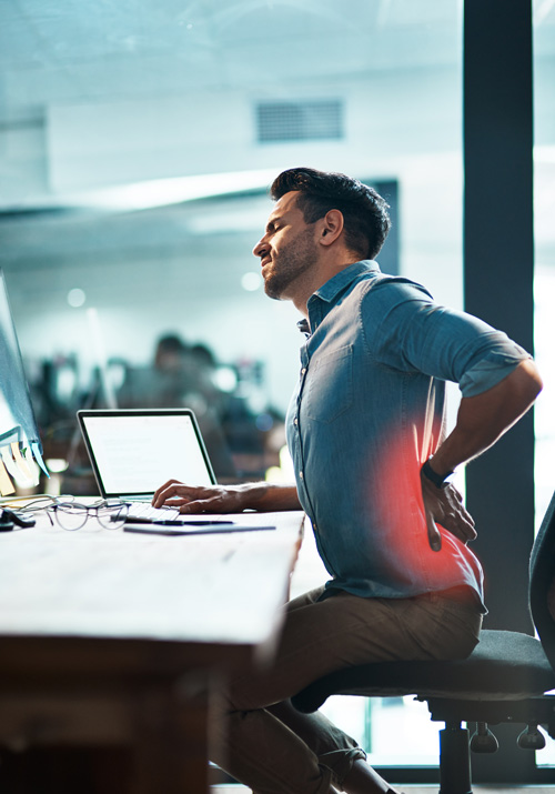 man sitting at a desk with back pain using a laptop in an office setting ergonomics for three steps to relief