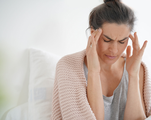 woman experiencing headache while sitting on a bed wearing a soft blanket feeling stressed and overwhelmed with pain relief in mind